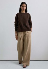 Sudadera de algodón marrón con hombros caídos, combinada con pantalones beige holgados y plisados. Un collar largo con un colgante ovalado completa el look.