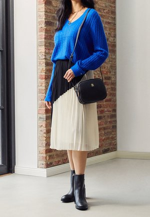 Blauw kabelgebreid truibloes, zwart-wit geplooide jurk, zwarte handtas en enkellaarzen. Gestructureerde materialen en contrasterende kleuren.