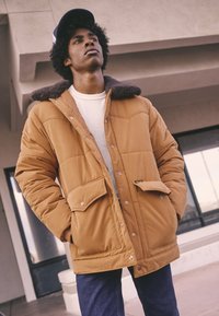 Casaco acolchoado castanho com colarinho texturado forrado a pelo, grandes bolsos frontais e fecho de pressão, usado sobre uma camisa branca de malha e jeans.