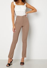 Body blanc à volants associé à un pantalon marron taille haute et cintré. Des talons ouverts noirs vernis complètent la tenue. Texture lisse dans l'ensemble.