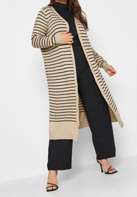 Béžový cardigan s černými horizontálními pruhy, dlouhými rukávy a zapínáním na knoflíky. Nošený přes černé tričko a spárovaný s černými kalhotami.