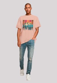 Helles rosa T-Shirt mit kurzen Ärmeln und einem Berg-Graphic, kombiniert mit verwaschenen blauen Jeans und weißen Turnschuhen.