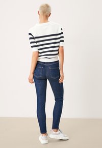 Pruhovaný svetr s krátkým rukávem v bílé a námořnické modři, v kombinaci s tmavě modrými skinny jeans a bílými teniskami s šedými akcenty, pohled zezadu.