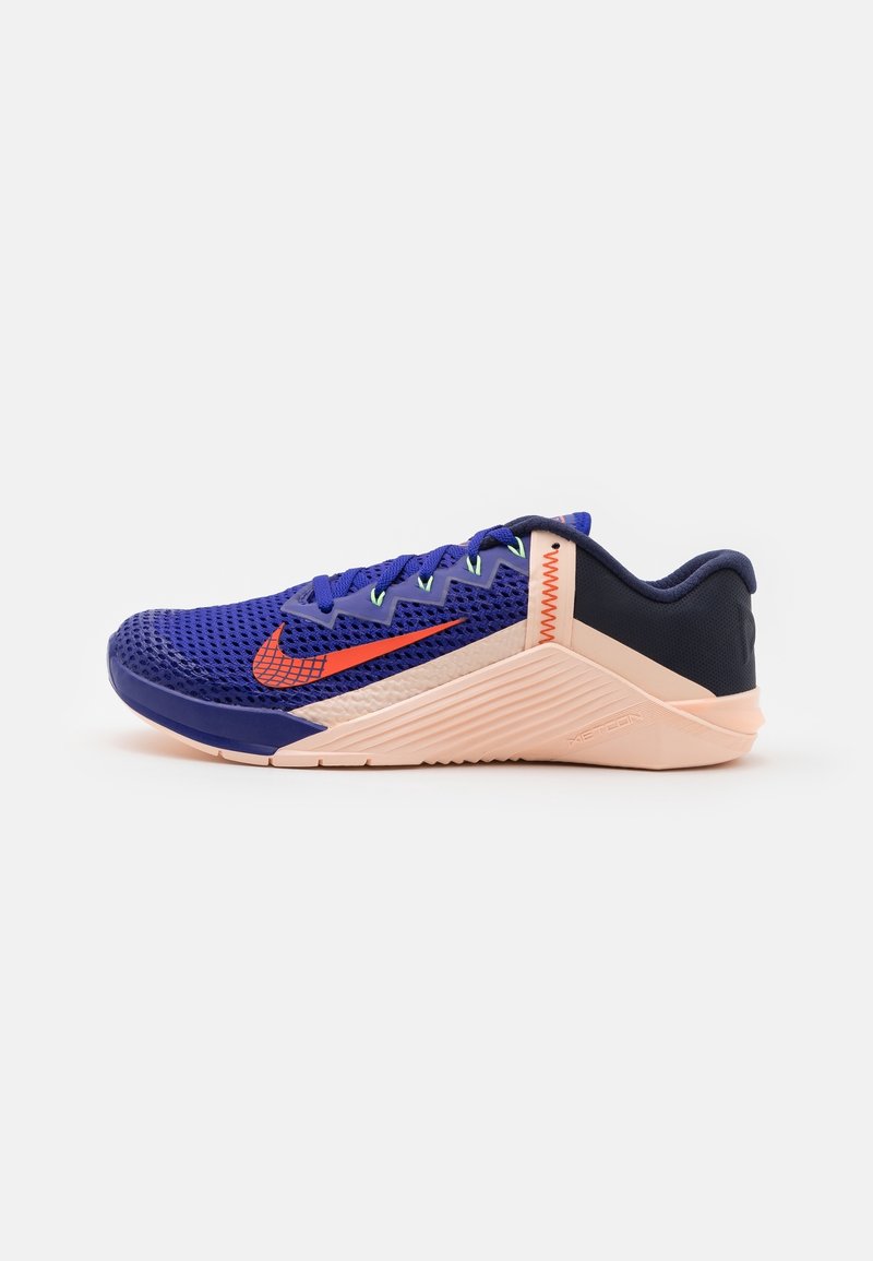 Scarpe da allenamento Nike con una tomaia in rete viola, swoosh arancione, dettagli navy e suola rosa chiaro. Presentano dettagli testurizzati e lacci.
