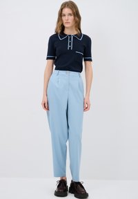 Marineblaues Poloshirt mit hellblauen Paspeln und Knopfdetail, kombiniert mit hellblauen hochtaillierten Hosen. Das Modell trägt dunkle Schnürschuhe.