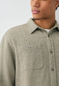 Light gray herringbone-patterned shirt with a button front, chest pocket, and collared neckline. Made from textured fabric.
