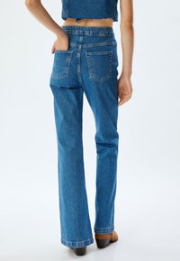 Jeans de denim azul acampanados con cintura alta, dos bolsillos traseros y un acabado limpio en los dobladillos. La textura de la tela es suave con un ligero desvanecimiento.