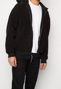 Person, der eine schwarze Fleecejacke mit Reißverschluss über einem weißen T-Shirt und schwarzer Hose trägt, vor einem einfarbigen hellen Hintergrund.