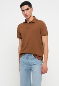 Καφέ πόλο μπλούζα με γιακά και κοντά μανίκια. Διαθέτει ένα μικρό λογότυπο στο στήθος, συνδυασμένη με ανοιχτό μπλε τζιν. Λείο υλικό.