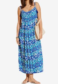 Vestido maxi azul con estampado, tirantes finos, escote fruncido y diseño de paisley. Modelo sosteniendo un bolso de mimbre y llevando sandalias planas.