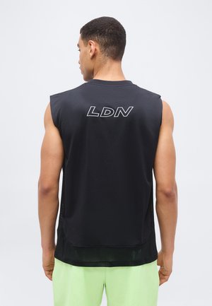 Ein Mann, von hinten gesehen, trägt ein ärmelloses schwarzes Shirt mit der Aufschrift "LDN" auf dem oberen Rücken und hellgrüne Shorts, vor einem einfarbigen Hintergrund.