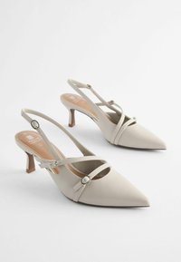 Kärmäkärkiset, beige slingback-korkokengät, joissa on ristinuoret ja hopeiset yksityiskohdat. Sileä nahkapinta, kohtuullinen korkeus ja minimalistinen muotoilu.