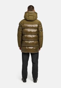 Olijfgroene pufferjas met een glanzende afwerking, voorzien van een capuchon, horizontaal gestikt patroon en opstaande kraag, gecombineerd met donkere broek.