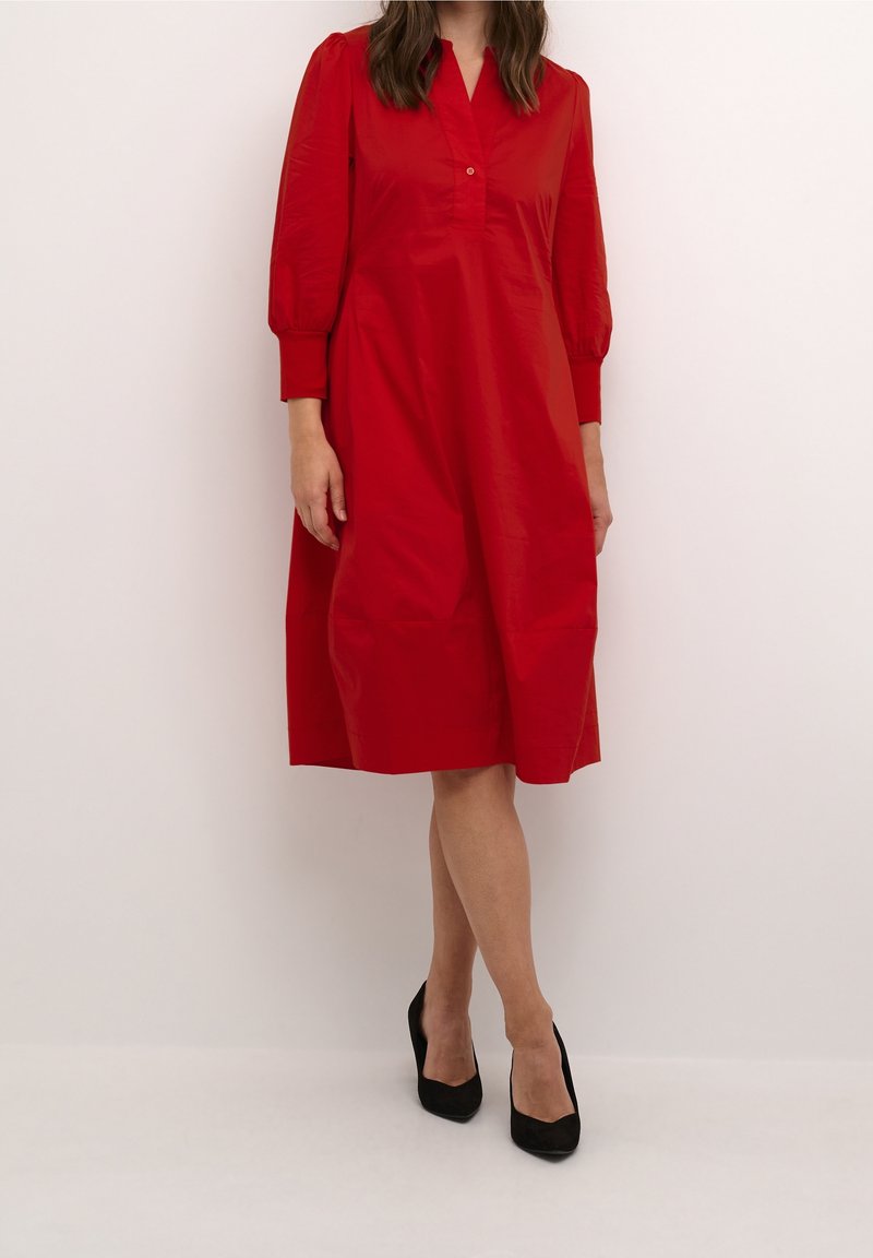 Abito rosso al ginocchio con scollatura a V, dettagli dei bottoni e maniche lunghe e puffate, realizzato in tessuto liscio. Abbinato a scarpe con tacco alto nere.
