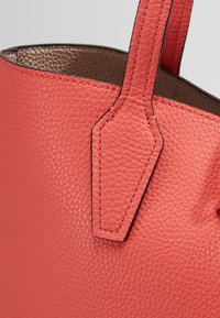Gros plan d'un sac à main en cuir texturé corail avec une sangle assortie et des coutures détaillées visibles.