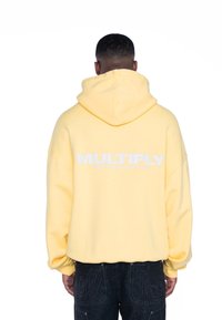 Sudadera amarilla de gran tamaño con un bolsillo canguro frontal y el texto blanco "MULTIPLY" en la parte posterior. Textura suave y diseño de corte relajado.