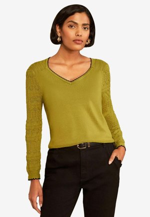 Woman wearing a green V-neck sweater with lace-patterned sleeves and black pants, standing with one hand in pocket against a white background.