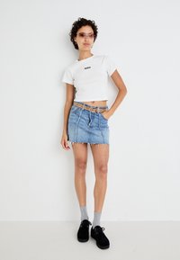 Haut court blanc avec logo noir, jupe en denim bleu clair avec ourlet effiloché, ceinture brune avec clous, chaussettes grises et chaussures à plateforme noires.