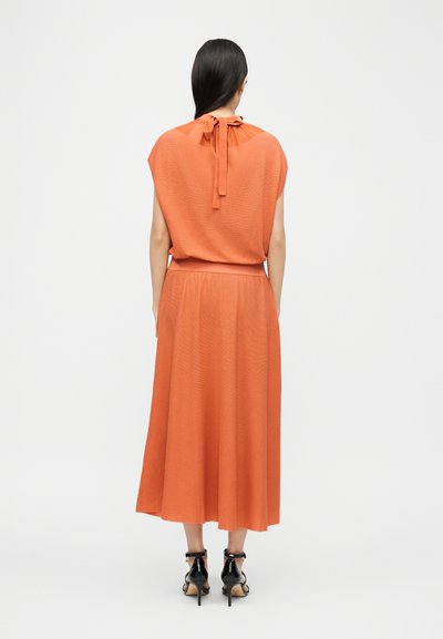 Femme aux longs cheveux foncés portant un haut sans manches orange avec un col noué et une jupe midi assortie, sandales à talons hautes noires, debout de dos.