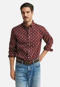 Homme aux cheveux foncés et à la moustache portant une chemise boutonnée bordeaux à motifs cachemire avec des manches retroussées, un jean bleu et une ceinture brune, se tenant devant un fond blanc.