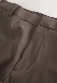 Gros plan de la ceinture avant d'un pantalon en tissu marron texturé avec passants pour ceinture et détail de poche cousue.