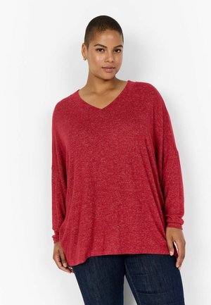 Maglietta rossa a maniche lunghe con scollo a V, caratterizzata da una vestibilità comoda e una texture morbida, abbinata a jeans scuri. Il tessuto presenta un motivo melangiato.