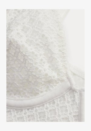 Valkoinen pitsikangas, jossa on kukkakuviot ja aaltoilevat reunat vaatekappaleen lähikuvassa.