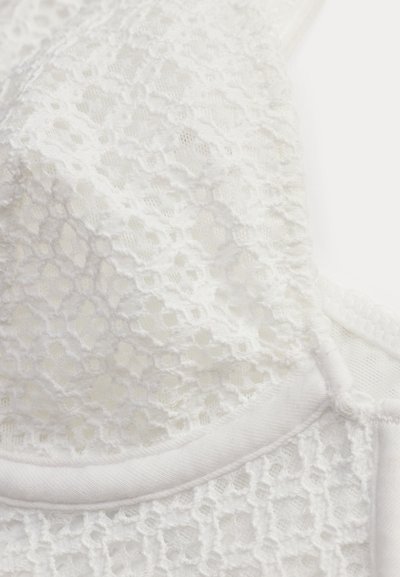 Λευκό δαντελένιο ύφασμα με λουλουδάτα σχέδια και κυματιστά άκρα σε κοντινή λήψη τμήματος ενός ρούχου.