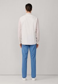 Witte mouwloze blouse over een lichtroze lange mouwen shirt, gecombineerd met lichtblauwe broeken en witte sneakers. Neutrale achtergrond.