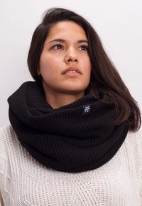 Black knitted infinity scarf with ribbed texture and a small logo tag, worn over a white cable-knit sweater. Soft and thick material.
