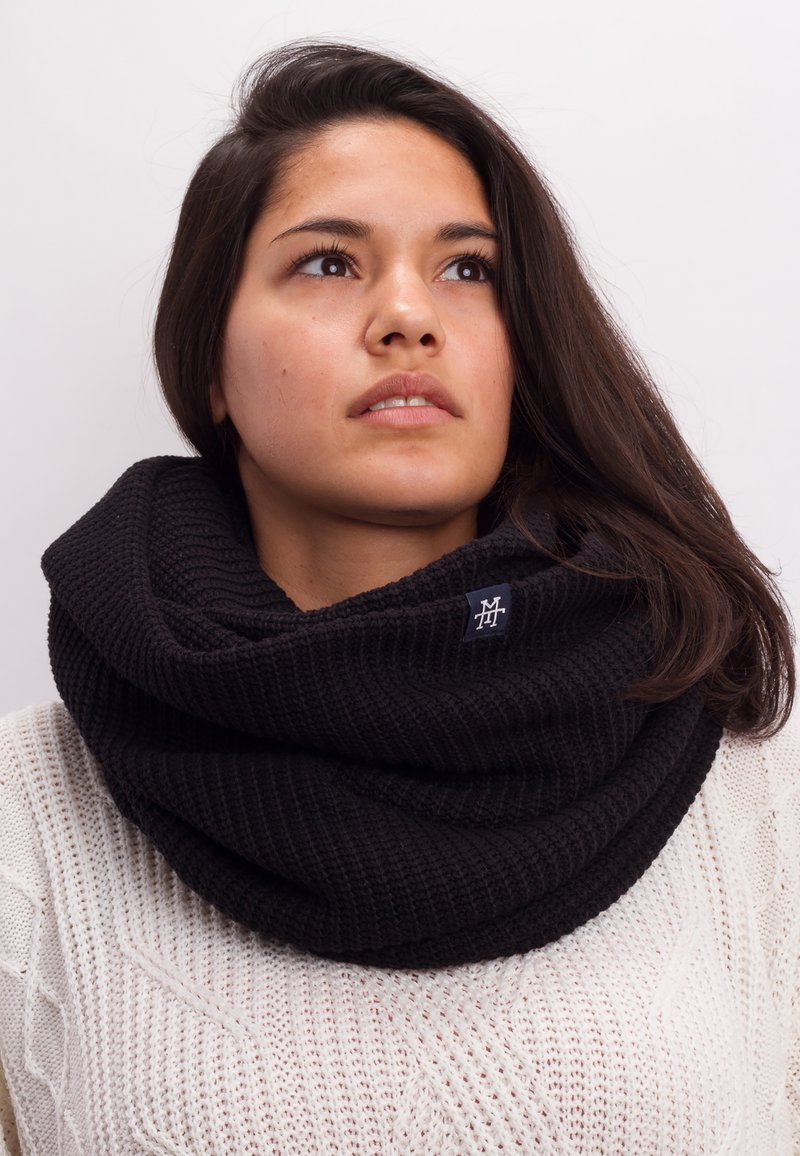 Black knitted infinity scarf with ribbed texture and a small logo tag, worn over a white cable-knit sweater. Soft and thick material.