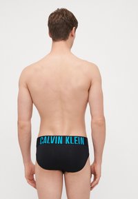 Schwarze Schwimmshorts mit einem breiten blauen Gummibund, der mit "CALVIN KLEIN" Branding versehen ist, im Rückansicht eines männlichen Modells.