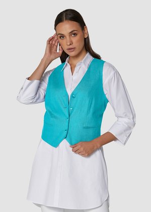Femme ajustant ses cheveux, portant un gilet boutonné turquoise vif sur une longue chemise blanche, sur fond uni.