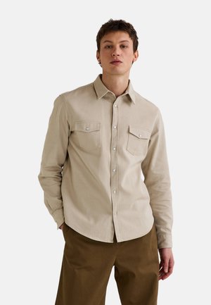 Chemise beige à manches longues en tissu texturé, dotée d'un devant boutonné, de deux poches poitrine et d'un ourlet arrondi. Associée avec un pantalon marron.