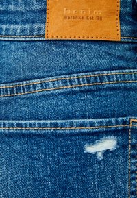 Pantalones vaqueros azul con un bolsillo trasero que tiene una etiqueta de cuero marrón con la inscripción "Denim Bershka Est. 98" y un ligero desgaste en la tela cerca del bolsillo.