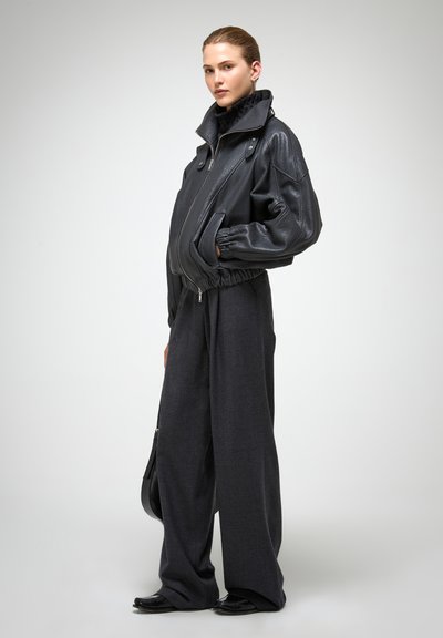 Schwarze Leder-Bomberjacke mit hohem Kragen, elastischen Bündchen und Reißverschluss vorne. Kombiniert mit locker sitzenden grauen Hosen und schwarzem Schuhwerk.