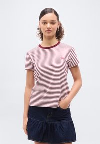 Νεαρή γυναίκα με κατσαρά μαλλιά, που φοράει κόκκινη και λευκή ριγέ μπλούζα Levi's και σκουρόχρωμη τζιν φούστα, στέκεται με το ένα χέρι στην τσέπη της.