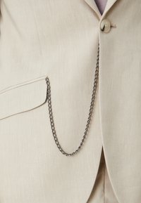 Beige blazer met een gestructureerd oppervlak, met een borstzak en een metallische kettingaccent. De ketting is zwart en gelinkt.