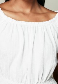 Blouse blanche texturée avec un col élastiqué froncé. Le tissu présente un motif de fronces subtil, créant du volume et de la douceur dans le design.