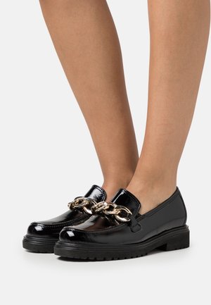 Mocassins en cuir verni noir ornés d'une chaîne dorée chunky, dotés d'une semelle plate et d'un bout rond. Texture lisse avec finition brillante.