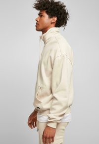 Beige høyhalset sweatshirt med frontlomme, ribbede mansjetter og avslappet passform. Har snor og myk tekstur.