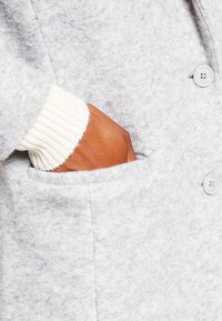 Abrigo de forro polar gris con un bolsillo, que presenta un puño blanco acanalado y grandes botones a juego. La textura parece suave y cálida.