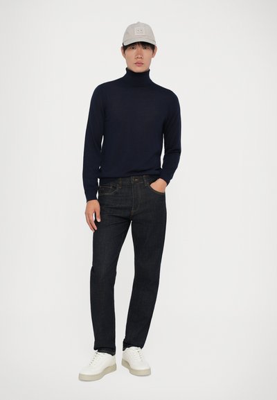 Marinefarbener Rollkragenpullover, dunkle Jeans und weiße Sneaker. Graue Mütze mit Logodetail. Glatte Textur und figurbetonter Schnitt.