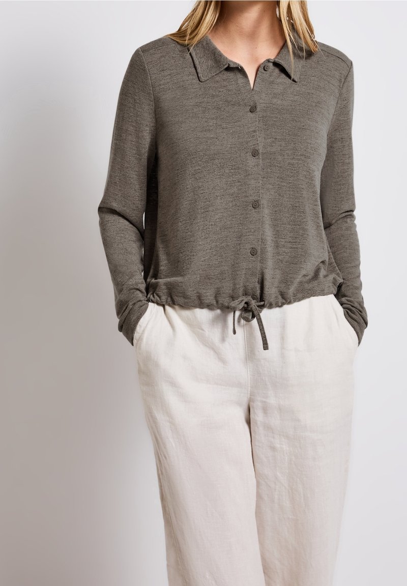 Haut vert à manches longues boutonné avec col et ourlet à cordon ajustable, porté avec un pantalon en lin crème léger.