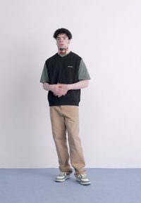 Čierny bezrukávový sveter cez zelené tričko, skombinovaný s béžovými nohavicami a teniskami s nádychom tyrkysovej. Svetlý pozadie; osoba stojí.