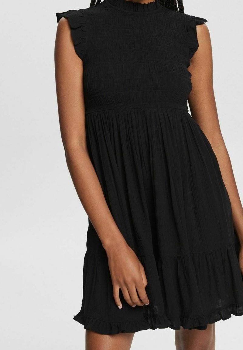 Femme portant une robe noire sans manches avec des épaules et un ourlet à volants, debout devant un fond uni gris clair.