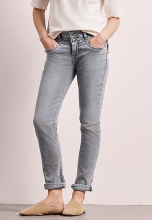 Person iført lysegrå slim-fit jeans med dobbelt knaplukning og beige slip-on sko, stående med hænderne i lommerne.