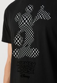 Černé tričko s potiskem Mickey Mouse ve stylu kostek a textem "MICKEY MOUSE" v tučném písmu na spodní části. Bavlněný materiál, krátké rukávy.