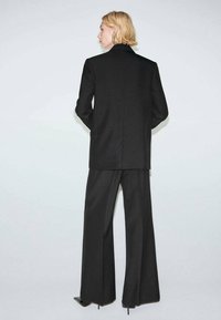 Czarna, dopasowana marynarka z wysokim kołnierzem, zestawiona z szerokimi spodniami o rozszerzanych nogawkach. Tkanina wydaje się strukturalna; posiada subtelną fakturę i ostre linie.