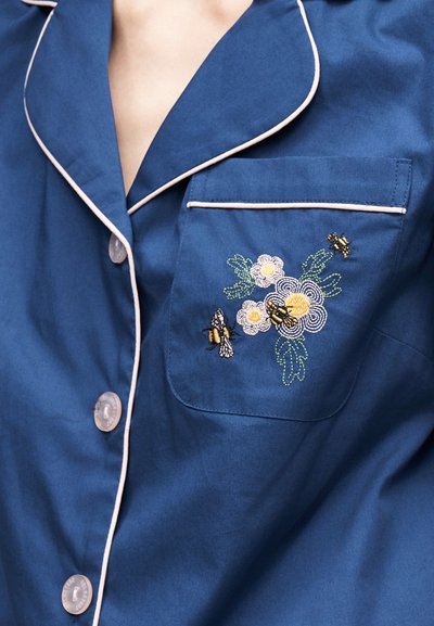 Μπλε παντελόνι πιτζάμας με λευκή διακόσμηση και κεντημένη τσέπη με λευκά λουλούδια και τρεις βουίζουσες μέλισσες.